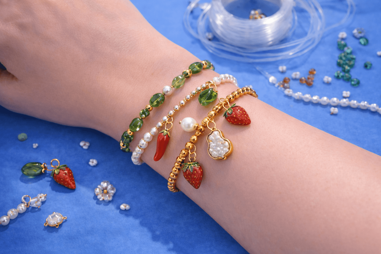Hand wearing stacked bracelets with strawberry and crystal charms on a blue surface